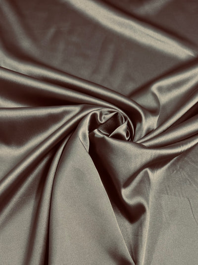 The Satin Deluxe - Pure Cashmere from Super Cheap Fabrics, 150cm wide, features smooth brown satin with a shiny finish that gathers softly in the center, creating luxurious folds and ripples as light gently enhances its texture.