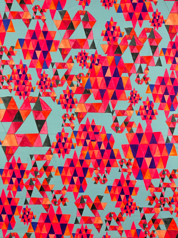 The "Geo Village" Printed Lycra from Super Cheap Fabrics showcases a complex, geometric pattern with numerous interconnected and overlapping triangles in shades of red, pink, orange, and purple against a light turquoise background. This visually engaging design measures 150cm wide and features vibrant hues and repetitive shapes that create a kaleidoscopic effect.