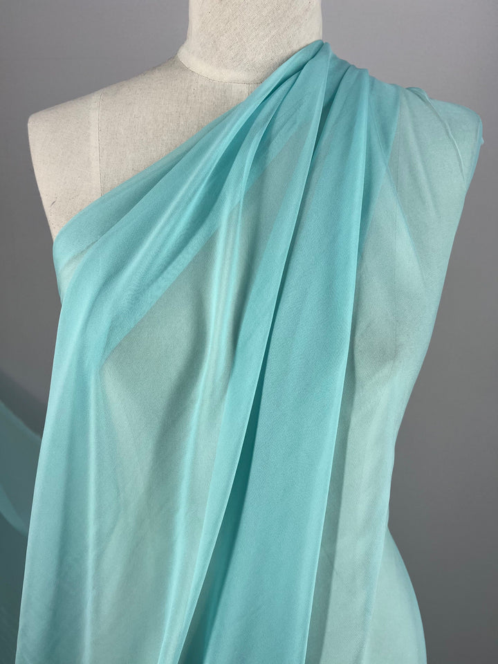 A mannequin is adorned in Super Cheap Fabrics' Silk Georgette - Tiffany - 135cm, gracefully draped over one shoulder to highlight the fabric's translucent and delicate qualities against a plain backdrop. This elegant look is ideal for spring and summer ensembles, capturing both grace and seasonal allure.