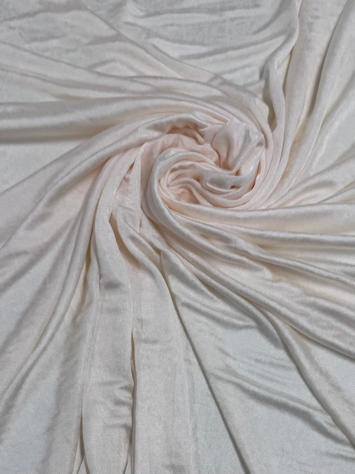 The Knit Lining Shimmy by Super Cheap Fabrics in cream features pale pink polyester artistically swirled at the center, forming a spiral pattern. Its lightweight, soft texture and subtle sheen capture the light beautifully. Width: 130cm.