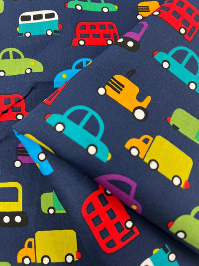 The "Printed Cotton - Mobile - 110cm" by Super Cheap Fabrics features a playful pattern of colorful vehicles, including cars, buses, and trucks on a dark blue backdrop. Made from natural cotton fibers in shades of red, yellow, green, blue, and purple, it's ideal for various uses.