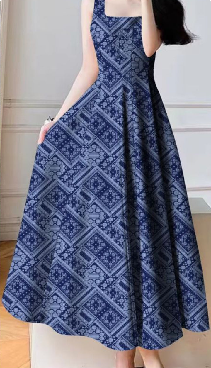 A woman stands indoors near a white wall and wooden floor, wearing a sleeveless, floor-length dress with a square neckline made from Super Cheap Fabrics' Designer Rayon - Navy Nomad - 145cm, featuring blue and white geometric and floral patterns.