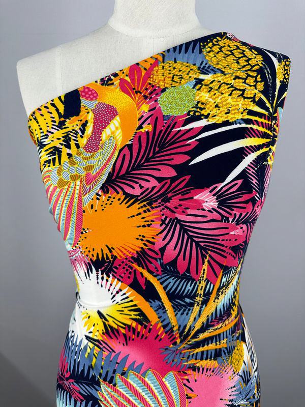 A mannequin is displaying a vibrant, asymmetrical one-shoulder dress crafted from Super Cheap Fabrics' Printed Lycra - Jumba, which measures 150cm. The dress features beautiful prints with an eye-catching tropical theme in colors like yellow, orange, pink, and black, showcasing palm leaves, flowers, and abstract patterns.