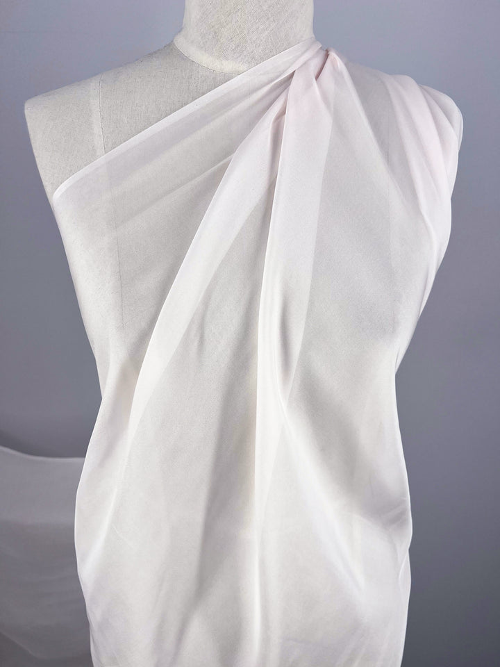 A mannequin draped in a piece of Pure Silk Chiffon - Bridal Blush from Super Cheap Fabrics, styled to mimic a toga-like garment. The extra light weight fabric is softly gathered at one shoulder, cascading down and creating gentle folds. The background is a plain, light gray wall.