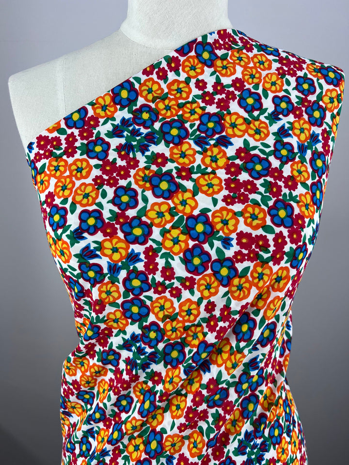 An elegant mannequin features a chic display of Super Cheap Fabrics' Salt Shrunk Print - Pansy - 155cm, showcasing a vibrant floral pattern with orange, blue, and red flowers amid green leaves on white. Styled asymmetrically across the shoulder, it exemplifies the allure of vibrant colors.