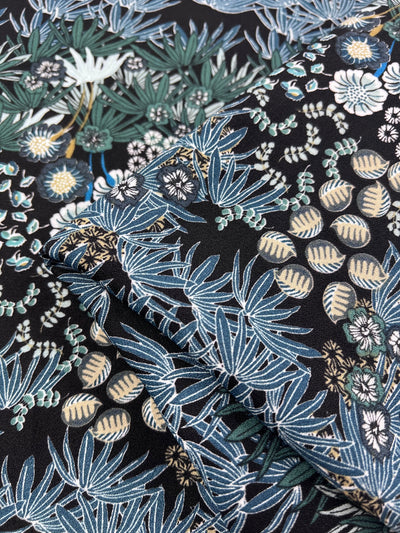 Close-up of Super Cheap Fabrics' Printed Crepe - Floral Reef, a lightweight polyester with intricate tropical patterns featuring blue, green, and beige leaves, elegantly paired with white flowers on a black background for a vibrant high-fashion look. Width: 150cm.