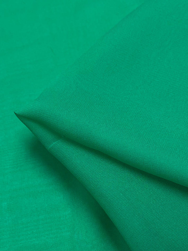 A close-up image of a smooth, emerald green Silk Georgette - 135cm fabric from Super Cheap Fabrics, slightly folded at one corner against a similar green background. The texture appears soft and even, with the fold creating subtle shadows and highlighting the material's flexibility.