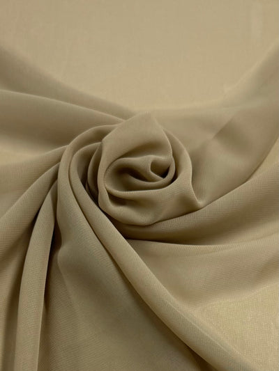 A close-up of Hi-Multi Chiffon - Tan 150cm by Super Cheap Fabrics shows the beige polyester fabric artfully arranged into a rose. The lightweight material forms natural folds and shadows, enhancing its texture and floral design.