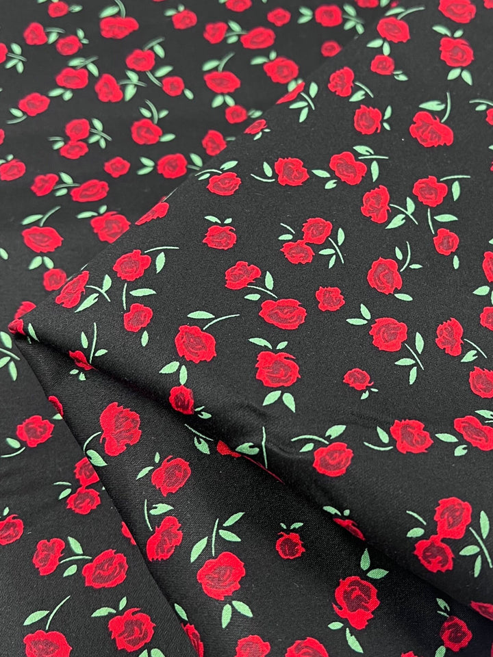 A close-up of Cotton Sateen - Rosely - 125cm by Super Cheap Fabrics reveals medium weight cotton with a repetitive pattern of red roses and green leaves on black, ideal for adding elegance to home décor with its smooth, folded appearance.