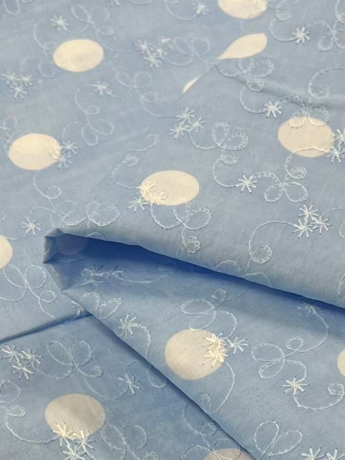 A close-up view of the "Embroidered Printed Cotton - Baby Blue - 112cm" from Super Cheap Fabrics showcases its natural cotton fibers in a light blue shade. The fabric is beautifully adorned with white embroidered patterns, highlighting large circles and delicate floral designs. Skillfully folded, it reveals different angles and textures, making it a perfect choice for both clothing and household items.