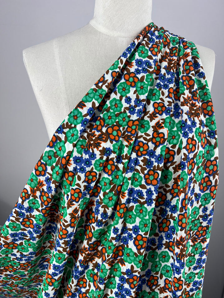 A mannequin displays Super Cheap Fabrics' Salt Shrunk Print - Kaleid, 155cm, featuring a vibrant floral design of orange, blue, and green flowers densely packed on white fabric, elegantly gathered over one shoulder.