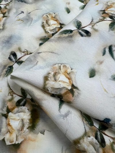A close-up of a piece of light-weight fabric showcases a delicate floral pattern. The design features soft, white roses and green leaves against a light, almost watercolor-like background, lending a serene and elegant feel to the material—ideal for children's clothing. This is Designer Cotton - Painted Roses - 145cm from Super Cheap Fabrics.