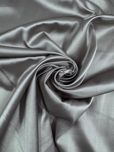 Close-up of Super Cheap Fabrics' Satin Deluxe in Satellite, 150cm, artfully twisted into a spiral. The glossy metallic gray satin reflects light softly off its folds, presenting a luxurious and elegant look.