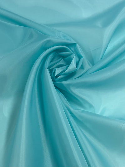 A close-up image of smooth, light blue satin fabric. The lightweight fabric is arranged in swirling folds, creating soft, gentle curves and a slightly glossy sheen that catches the light, highlighting its silky texture. This is the Lining - Angel Blue - 150cm by Super Cheap Fabrics.