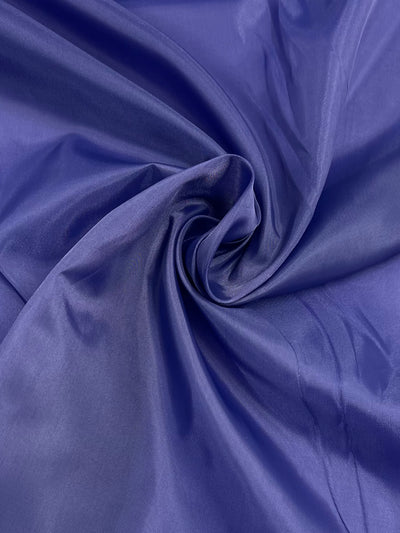 A close-up view of a soft, deep blue silk Super Cheap Fabrics Lining - Purple - 150cm with a smooth and shiny texture. The fabric is artistically folded and gathered in the center, creating a swirling pattern that accentuates its luxurious sheen, perfect for elegant garment accents.