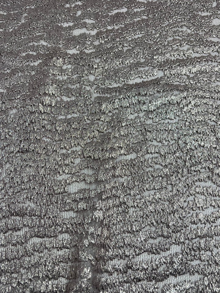 Close-up of a metallic silver textured fabric with a pattern of irregular, wavy lines creating a shimmering and reflective surface. This polyester lace fabric, Evening Lace - Avalanche - 150cm by Super Cheap Fabrics, captures light, producing subtle variations in shade and sheen.