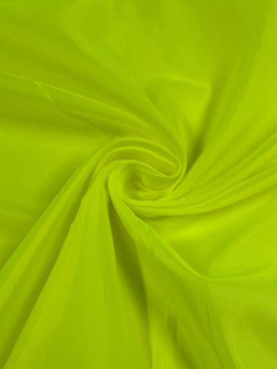 A close-up of a bright neon yellow lightweight fabric, twisted in the center creating subtle folds and patterns. The smooth, shiny texture of the vibrant fabric is visible, highlighting its soft appearance. This fabric is called Lining - Lime - 150cm by Super Cheap Fabrics.