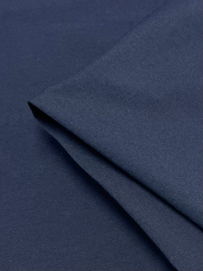 Close-up image of a dark blue, medium-weight fabric, showing its smooth texture and slight fold, suggesting a soft and flexible material. The fabric appears to be neatly draped, highlighting its even color and fine weave, typical of high-quality Cotton Lycra - Navy - 150cm from Super Cheap Fabrics.