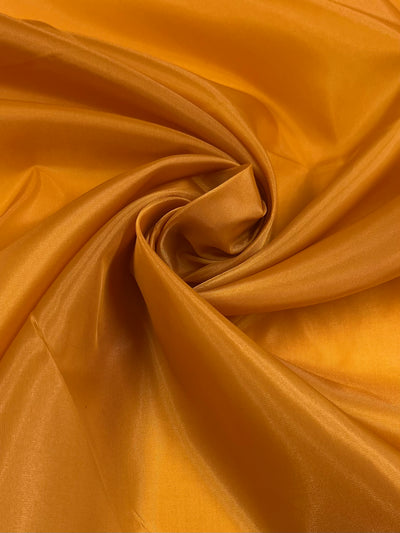 A close-up of a piece of glossy, golden-yellow polyester lining. The vibrant fabric is arranged in a swirling pattern, creating a sense of texture and depth with its shiny, smooth surface reflecting light. The product shown is Lining - Dark Cheddar - 150cm from Super Cheap Fabrics.