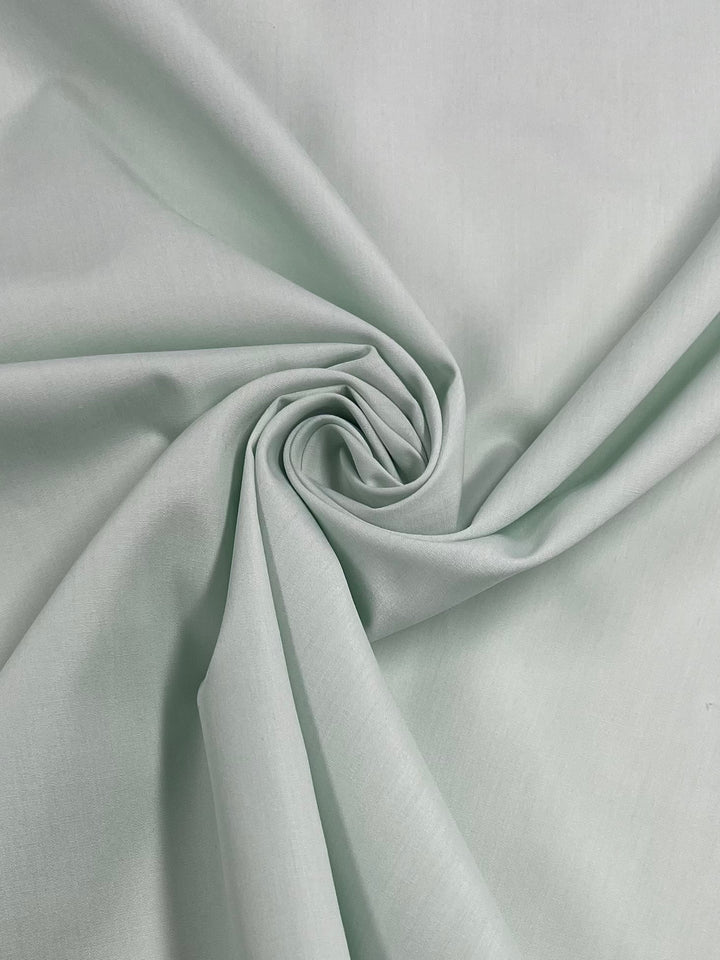 A close-up of the Poplin - Ice Green fabric by Super Cheap Fabrics reveals its smooth texture, crafted from a polyester-cotton blend. The light green material is gathered in a swirl pattern at the center, with its lightweight and soft folds creating a visually appealing spiral shape that highlights its elegant drape.