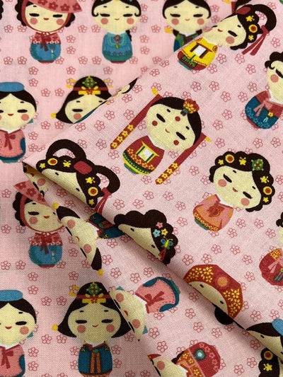 Super Cheap Fabrics' Printed Cotton - Authentic Pink - 112cm features a repetitive pattern of cute, cartoonish characters dressed in traditional Asian attire. The characters, depicted with various styles and colors, have cheerful expressions and are adorned with flowers and accessories—perfect for quilting or other crafts.