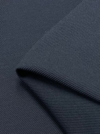 Close-up view of a folded piece of the Stretch Wool Twill - Midnight- 125cm from Super Cheap Fabrics. The dark gray fabric showcases a textured surface with a visible weave pattern, featuring fine diagonal lines that add depth and detail to this medium-weight material. This Italian fabric appears smooth and slightly glossy under the light.