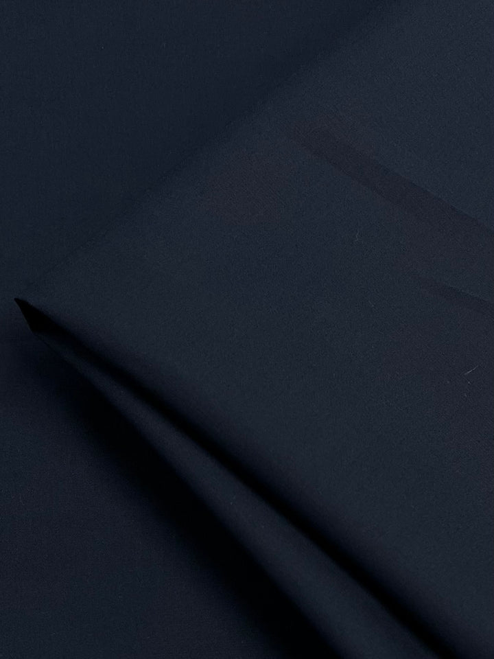 A close-up of a smooth, dark navy blue fabric folded over itself. The 100% cotton surface appears soft and slightly reflective, with gentle creases where the lightweight fabric is layered. This is the Cotton Voile - Navy - 145cm by Super Cheap Fabrics.