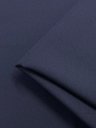 A close-up of a folded piece of Crepe Suiting - Navy - 145cm from Super Cheap Fabrics, showing its texture and weave. The 100% polyester material appears smooth and tightly woven, with a slight sheen. The image focuses on the corner fold, highlighting the medium weight fabric's finish and quality.