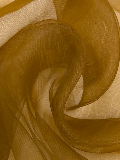 A close-up of Super Cheap Fabrics' Organza - Meerkat - 150cm reveals a swirling motion showcasing its rich, golden-brown hue. The lightweight fabric's delicate texture and translucent quality emphasize movement and fluidity, with light subtly highlighting its graceful curves.