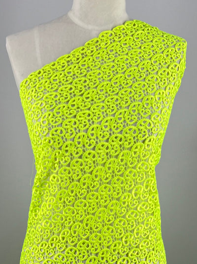 A mannequin draped in vibrant sharp green Anglaise Lace from Super Cheap Fabrics, often used in bridal wear, featuring an intricate chain-link pattern. The fabric is wrapped asymmetrically across the mannequin, covering one shoulder and leaving the other bare. The plain gray background highlights the bright lace fabric perfectly.