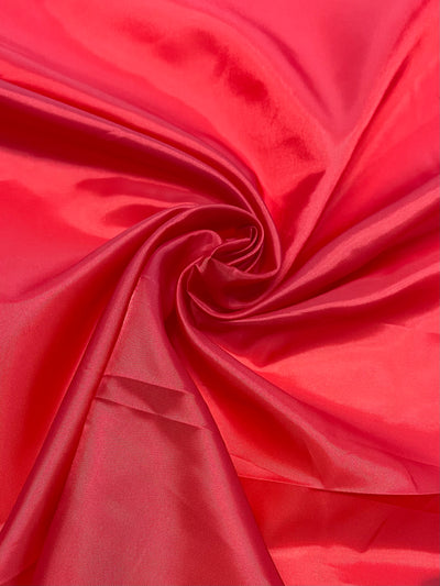 A close-up of vibrant Lining - Red - 120cm from Super Cheap Fabrics reveals a soft, smooth texture. The lightweight fabric is gathered in the center, creating graceful, swirling folds that catch the light and highlight its glossy, luxurious surface. Subtle hints of polyester lining add to its durability and elegance.