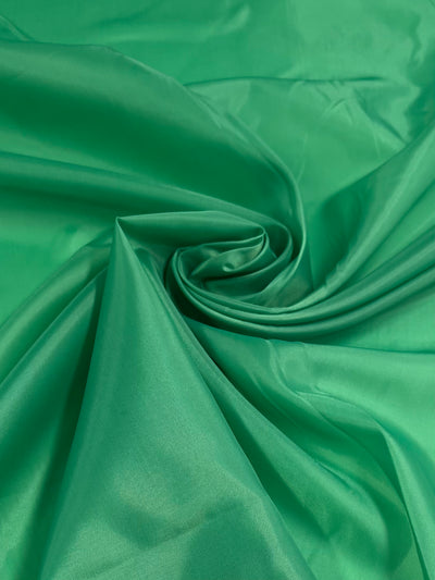 A close-up of smooth, Lining - Emerald Green - 150cm from Super Cheap Fabrics, artistically arranged and swirling into a central point, creating a spiral effect. The polyester lining appears glossy and has a silky texture.