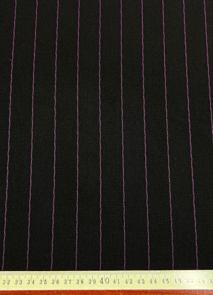 The Microfibre Suiting - Black/Pink Stripe - 150cm by Super Cheap Fabrics is displayed, featuring a medium-weight black polyester fabric adorned with uniformly spaced, thin pink pinstripes running vertically. A measuring tape at the bottom edge shows dimensions in centimeters, indicating a fabric width of approximately 150 centimeters.