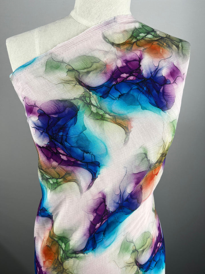 A mannequin is adorned with the Designer Cotton - Azurine fabric from Super Cheap Fabrics, which showcases a vibrant abstract pattern. The design features swirling shades of blue, purple, green, and orange that evoke watercolor or smoke effects on a light background, making it ideal for luxury sewing projects using premium designer cotton fabrics.