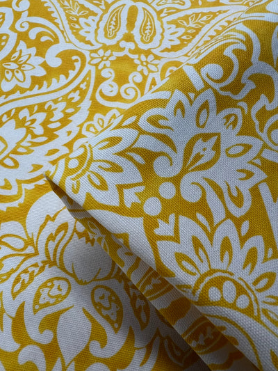 A close-up image of Super Cheap Fabrics' Printed Duck Canvas - Damask - 145cm with a white and mustard yellow floral and paisley pattern. The intricate design, perfect for home décor or fashion accessories, features various shapes and curves. The fabric is slightly folded in one corner, creating a layered look.
