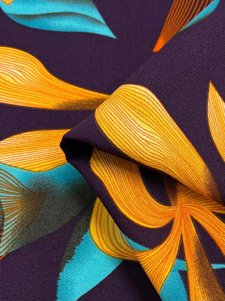 Close-up of a vibrant polyester fabric with a floral pattern. The design features large, stylized flowers with yellow petals and blue-green leaves on a dark purple background. The lightweight fabric is folded, creating dynamic curves and shadows, perfect for high fashion creations. This is the Printed Crepe - Wild Lily - 150cm from Super Cheap Fabrics.