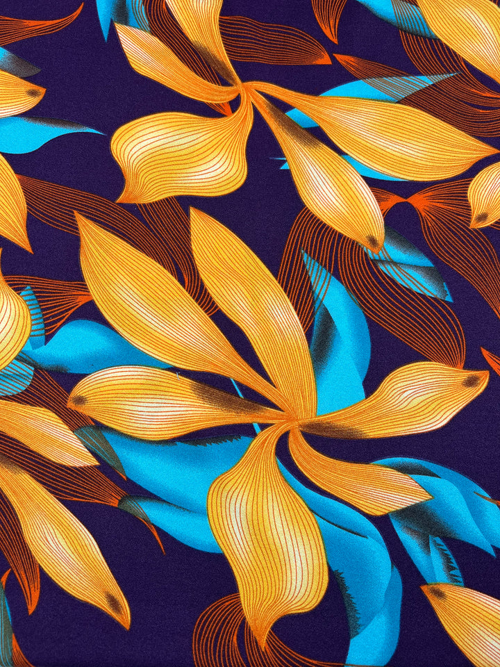 A vibrant polyester fabric pattern featuring large, abstract golden-yellow and teal leaves with fine lines, set against a dark blue-purple background. The leaves are interwoven, creating a dynamic and flowing design ideal for high fashion pieces. Product Name: **Printed Crepe - Wild Lily - 150cm** Brand Name: **Super Cheap Fabrics**