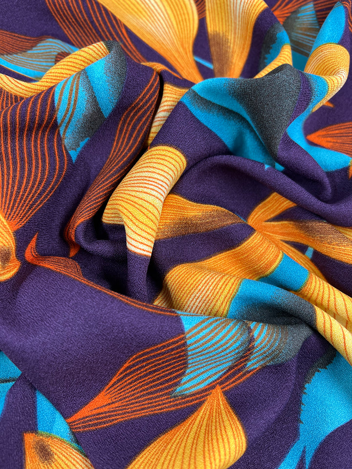 A close-up of a high fashion polyester fabric featuring an abstract pattern with vibrant colors. The design includes flowing shapes in shades of orange, blue, yellow, and teal on a deep purple background, creating a sense of movement and texture. The lightweight fabric appears crumpled. This is the Printed Crepe - Wild Lily - 150cm by Super Cheap Fabrics.