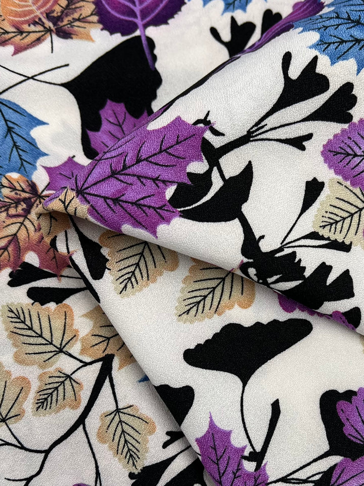 Close-up of Super Cheap Fabrics' Printed Crepe - Colourful Fall - 150cm featuring a vibrant autumn leaf pattern. The high fashion design includes black, purple, blue, and tan leaves on a white background, with various leaf shapes overlapping each other, creating an intricate and colorful texture.