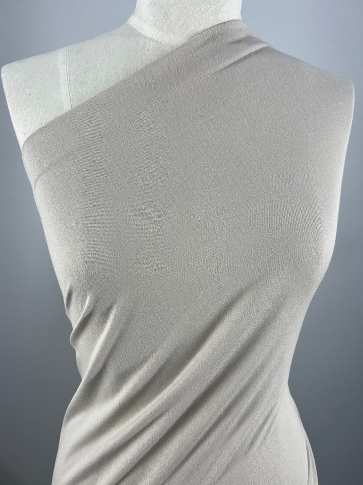A white mannequin is draped in a beige, one-shoulder, 100% cotton fabric called Cotton Jersey - Oatmeal - 145cm from Super Cheap Fabrics. The material appears smooth and form-fitting, showcasing a minimalistic, elegant design. The background is a plain gray.