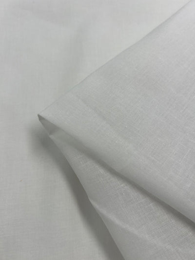 A close-up of a neatly folded piece of plain white fabric lying on an unfolded section of the same material. This 100% cotton, extra light weight fabric has a smooth and crisp texture, and the image's lighting emphasizes its clean and uniform appearance, perfect for home decor projects. **Super Cheap Fabrics' Cotton Voile - White - 145cm** is particularly well-suited for such applications.