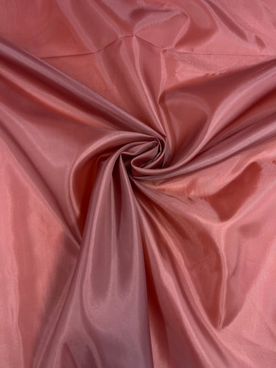A close-up image of **Super Cheap Fabrics' Lining - Tandoori Spice - 150cm**, a soft, silky, lightweight lining fabric in a pinkish-brown color, artistically gathered and twisted into a rosette-like shape in the center, creating an elegant and smooth texture with subtle folds and a slight sheen.
