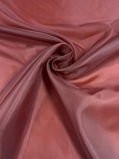 Close-up of a piece of smooth, shiny Super Cheap Fabrics Lining - Bossa Nova - 150cm in a rich, reddish-pink color. The fabric is arranged with folds that converge into a swirl pattern in the center, creating a sense of motion and texture. The surface reflects light, highlighting its glossy finish.