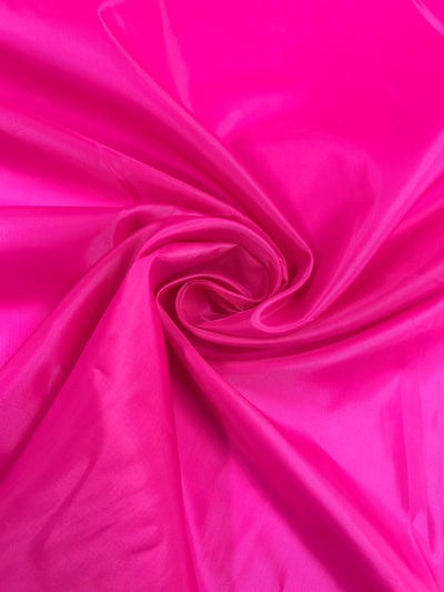 A close-up image of a vibrant pink fabric, twisted and arranged to form a swirling pattern in the center. The texture appears smooth and slightly shiny, reflecting light to create variations in the shade of pink. This Super Cheap Fabrics Lining - Hot Pink - 150cm adds an elegant touch with its lightweight feel.