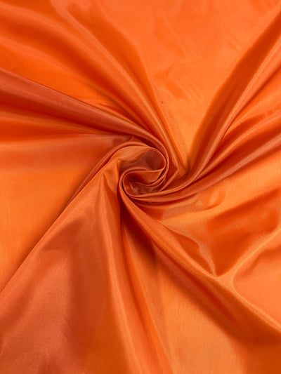 A close-up shot of vibrant orange polyester lining fabric with a smooth, shiny texture. The lightweight Super Cheap Fabrics Lining - Tigerlily - 150cm is gently twisted from the center, creating soft folds and a sense of depth. The lighting highlights the fabric's rich color and sheen perfectly.