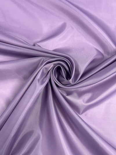 A close-up of smooth, glossy Orchid Mist - 150cm fabric from Super Cheap Fabrics swirled into a spiral pattern. The polyester lining has a shiny finish, creating highlights and shadows that emphasize its silky texture.