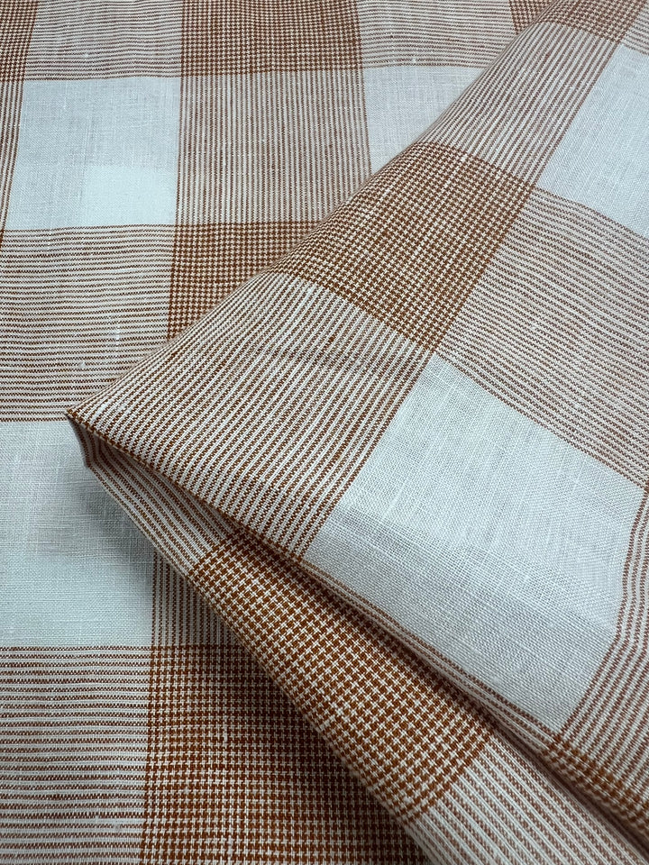 A close-up of a folded lightweight fabric with a large orange and white checkered pattern. Each square contains a combination of solid and striped designs, creating a textured appearance. The fabric appears to be made of woven pure linen, enhancing its delicate charm. This is the Pure Linen - Copper Check - 145cm by Super Cheap Fabrics.