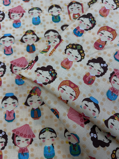 This 100% cotton quilting fabric showcases a colorful pattern of cartoon girls in various traditional Asian outfits and hairstyles. With round faces, closed eyes, and adorned with accessories like flowers, hats, and hairpieces, this elegant fabric is perfect for childrenswear. The background is a light beige color. The product is called Printed Cotton - Authentic - 112cm from Super Cheap Fabrics.