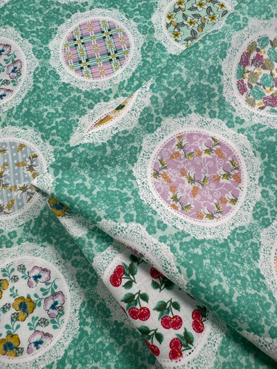 A fabric with a green background features circular patches of various patterns, including plaid, cherries, flowers, and butterflies, all bordered with lace. Perfect for quilting and other crafts, this 100% cotton material is slightly folded at the center of the image. Introducing Printed Cotton - Mixed Mirrors - 112cm by Super Cheap Fabrics.