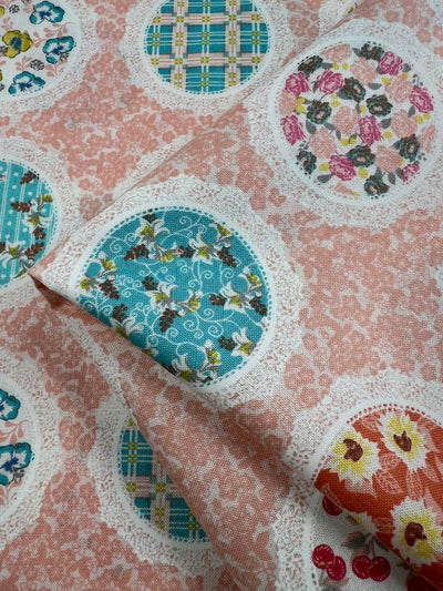 A piece of Super Cheap Fabrics' Printed Cotton - Pink Mixed Mirrors - 112cm with a pink background features various circular patterns. Each circle contains distinct floral and checkered designs in colors like blue, green, and red. The cotton homespun fabric is slightly folded, displaying the different patterns prominently.